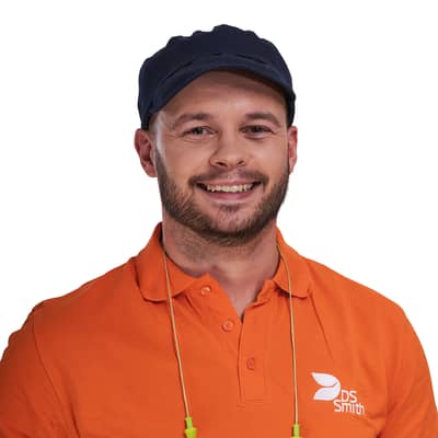 Portrait eines jungen Mannes, Bart, Mütze und Gehörschutz mit orangenem T-Shirt, Arbeitskleidung der Produktion. Er lächelt in die Kamera.