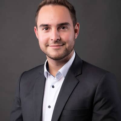 Portrait eines jungen Mannes mit kurzen, dunklen Haare. Weißes Hemd, dunkles Jacket. Er schaut in die Kamera. 