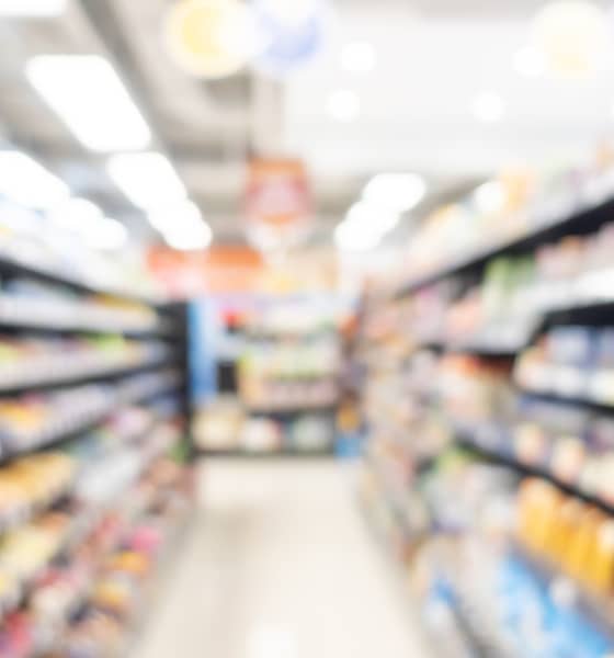 Verschwommener Gang eines Supermarkts mit vollen Regalen rechts und links sowie hinten im Bild.