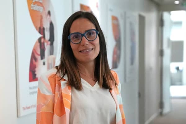 Portrait de Jessica Truchet dans un couloir de bureau, portant des lunettes bleues, un chemisier blanc et une veste à motifs orange.