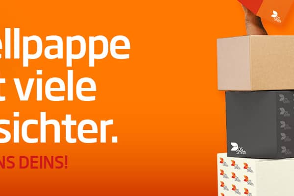 dunkelhaariger junger Mann steht rechts neben und halb hinter einem Kistenstapel aus Wellpappe, freundlich in die Kamera lächelnd, mit einem kleinen Wellpappkarton in der Hand. Mütze und Gehörschutz und oranges T-Shirt, Arbeitskleidung, vor orangefarbenem Hintergrund