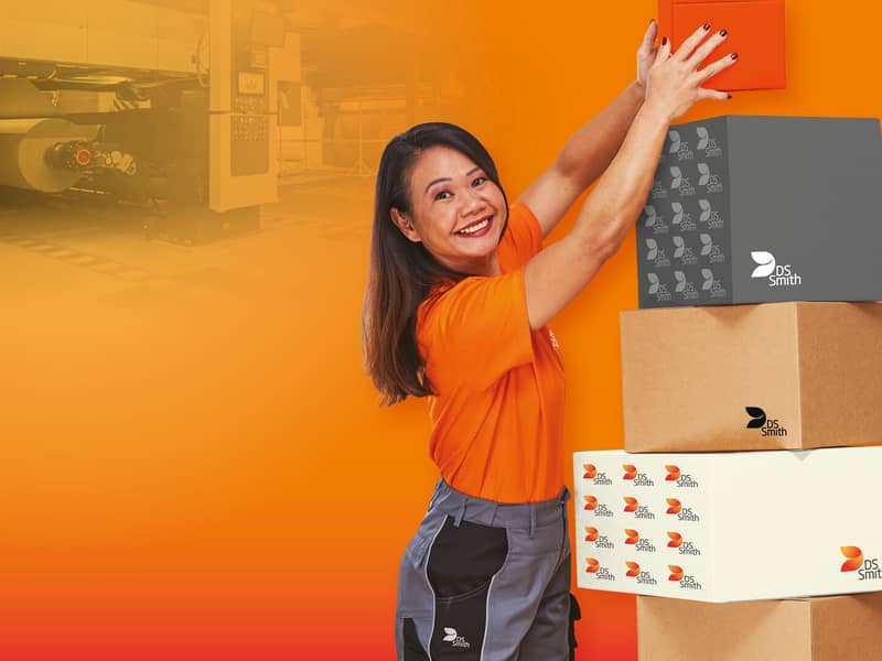 asiatische Frau mit langen Haaren steht links eines Kartonstapels aus Wellpappe vor orangefarbenem Hintergrund und lächelt in die Kamera. Sie trägt ein orangefarbenes T-Shirt was deutlich Arbeitskleidung der Produktion ist. Sie hält mit beiden Händen einen kleinen Karton aus Wellpappe und stellt diesen auf den Stapel oben drauf.