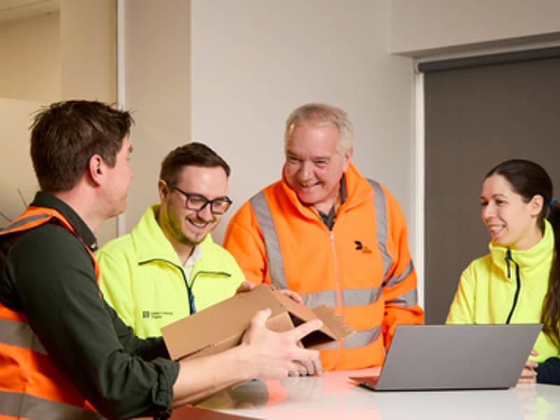 Four International Paper and DS Smith workers in reflective vests talk in front of a laptop. One of them holds an item of cardboard packaging.