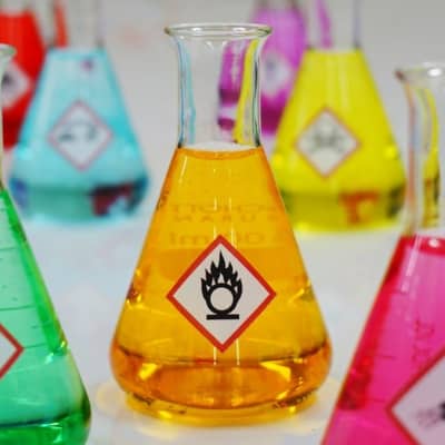 Mehrere Laborgläser gefüllt mit Gefahrgutetiketten und bunten Flüssigkeiten.