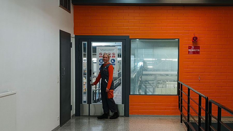 Naishenkilö oranssi-harmaissa haalareissa avaamassa ovea aaltopahvitehtaan tuotantoon