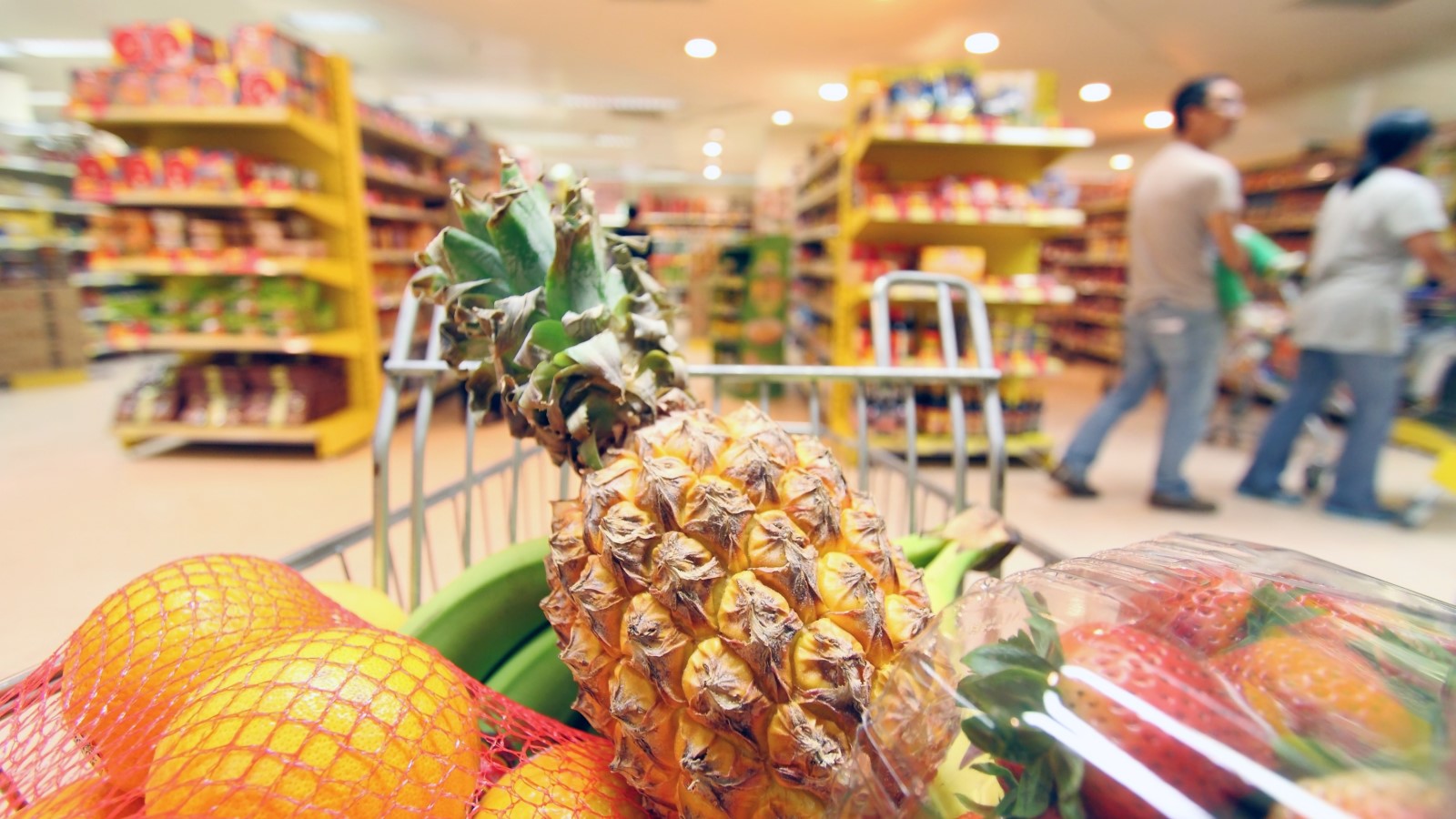 Eine Ananas im vollen Einkaufswagen ist fokussiert, während im Hintergrund ein Supermarkt zu sehen ist. 