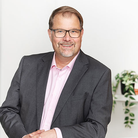 kuvassa Ari Viinikkala niminen mies hymyilee ja katsoo kameraa harmaa puku päällään nojaten pöytään