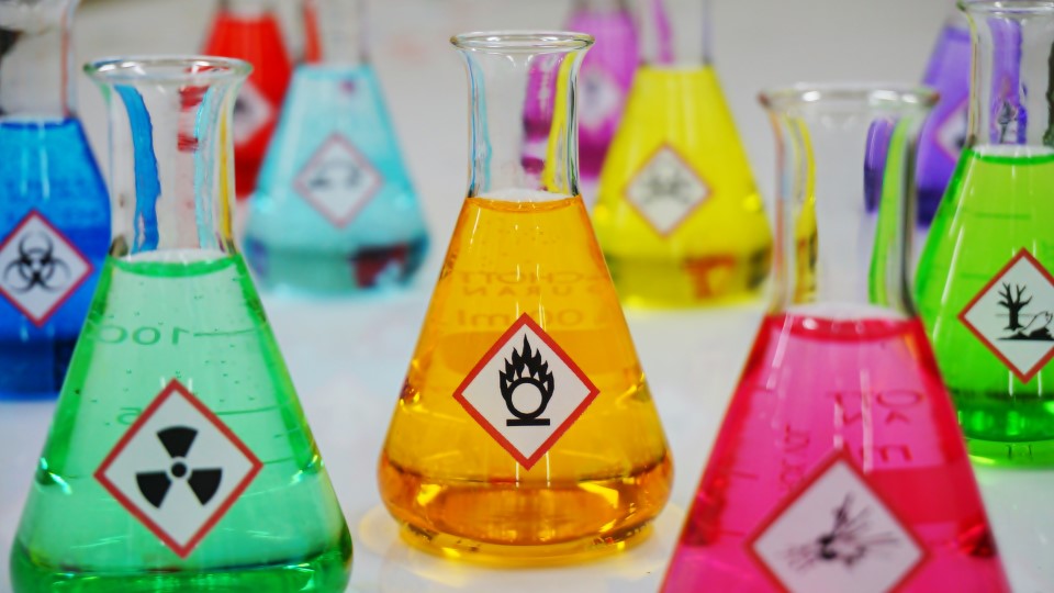 Mehrere Laborgläser gefüllt mit Gefahrgutetiketten und bunten Flüssigkeiten.