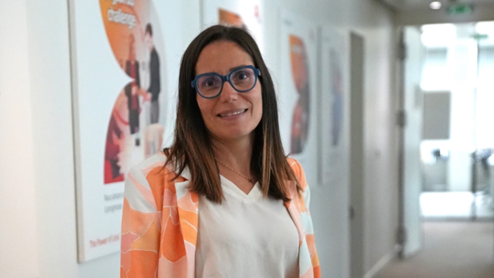 Portrait de Jessica Truchet dans un couloir de bureau, portant des lunettes bleues, un chemisier blanc et une veste à motifs orange.