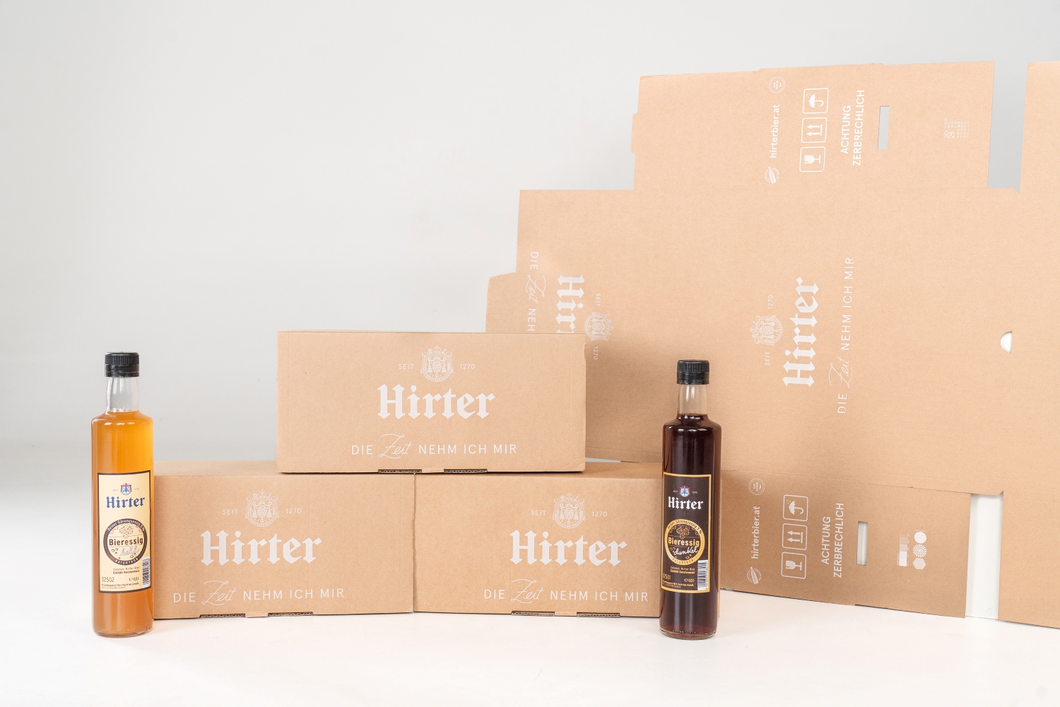 Gesamtansicht der neuen Verpackungslösung von DS Smith für die Privatbrauerei Hirt. Im Bild sind drei zusammengefaltete Verpackungen, ein ungefalteter Bogen stehend und zwei Flaschen mit Hirter Bieressig.