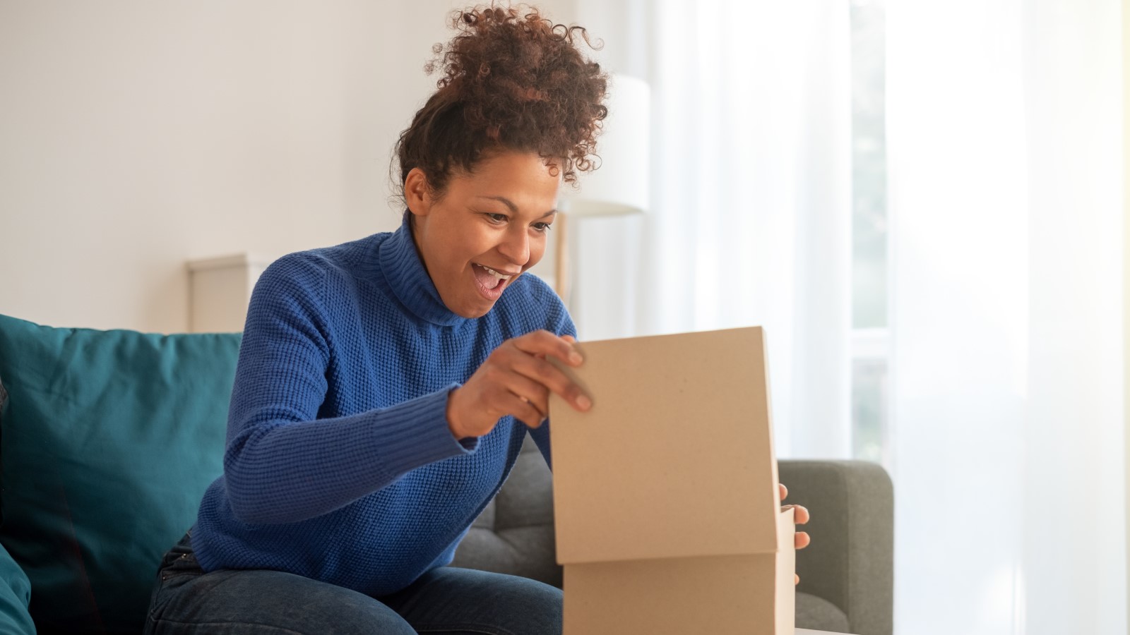 Eine Frau sitzt auf dem Sofa und freut sich, während sie ein Paket öffnet.