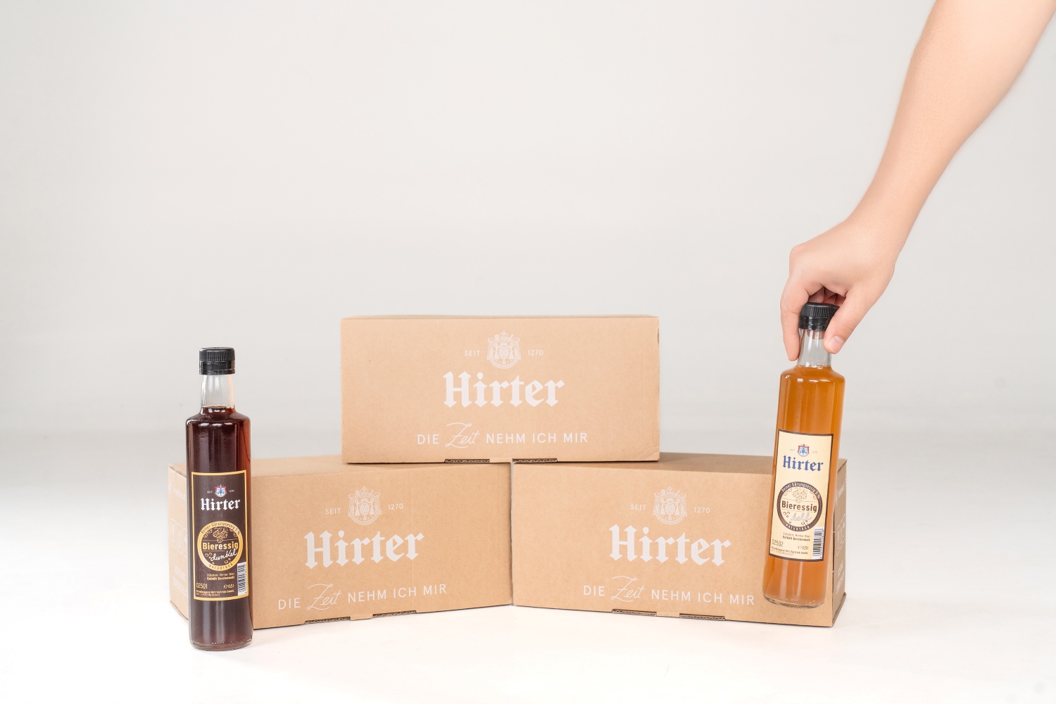 Ansicht der neuen Verpackungslösung mit zwei Hirter Essigflaschen. Eine Hand greift nach einer der Flaschen.