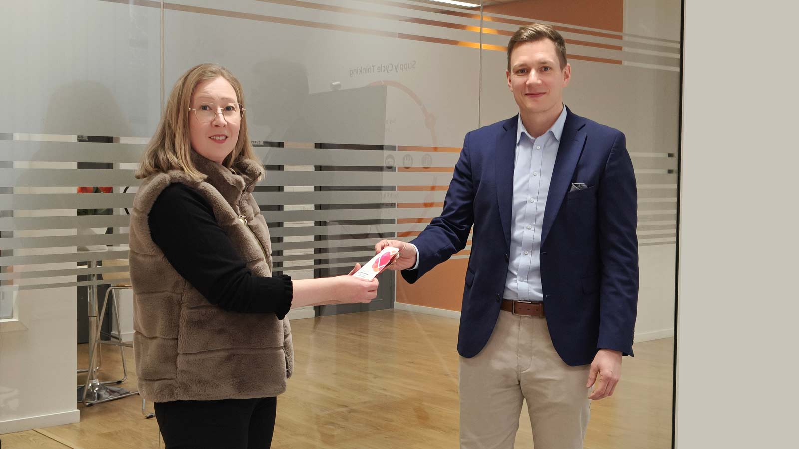 Kuvassa nainen ja mies, ja mies ojentaa naiselle Auttamisesta arkea järjestön lahjoituskortin, jolla nainen voi ohjata miehen antamat rahat haluamalleen hyväntekeväisyyskohteelle