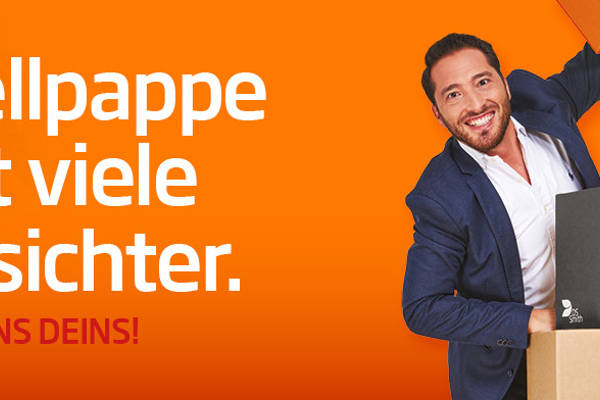 junger dunkelhaariger Mann mit dunklem Jacket steht links hinter einem Wellpappaufsteller und hält ein Wellpappkarton in der Hand und lächelt in die Kamera.