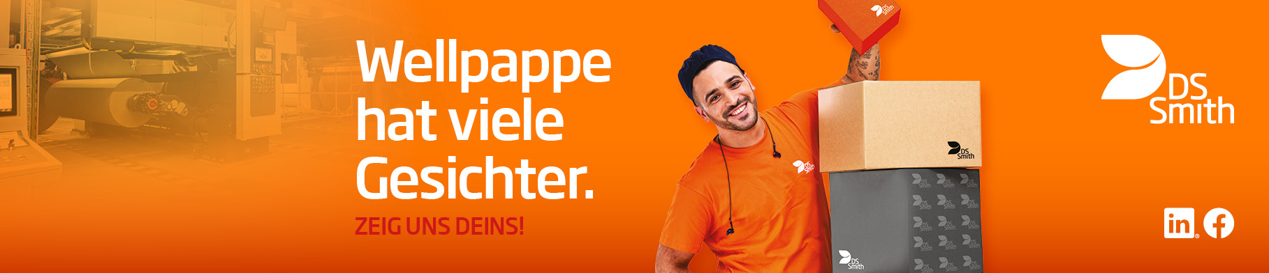  dunkelhaariger junger Mann steht links neben einem Kistenstapel aus Wellpappe, freundlich in die Kamera lächelnd, mit einem kleinen Wellpappkarton in der Hand. Mütze und Gehörschutz und oranges T-Shirt, Arbeitskleidung, vor orangefarbenem Hintergrund