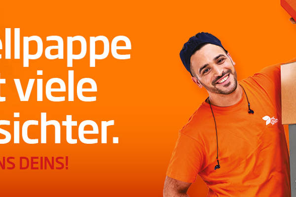 dunkelhaariger junger Mann steht links neben einem Kistenstapel aus Wellpappe, freundlich in die Kamera lächelnd, mit einem kleinen Wellpappkarton in der Hand. Mütze und Gehörschutz und oranges T-Shirt, Arbeitskleidung, vor orangefarbenem Hintergrund
