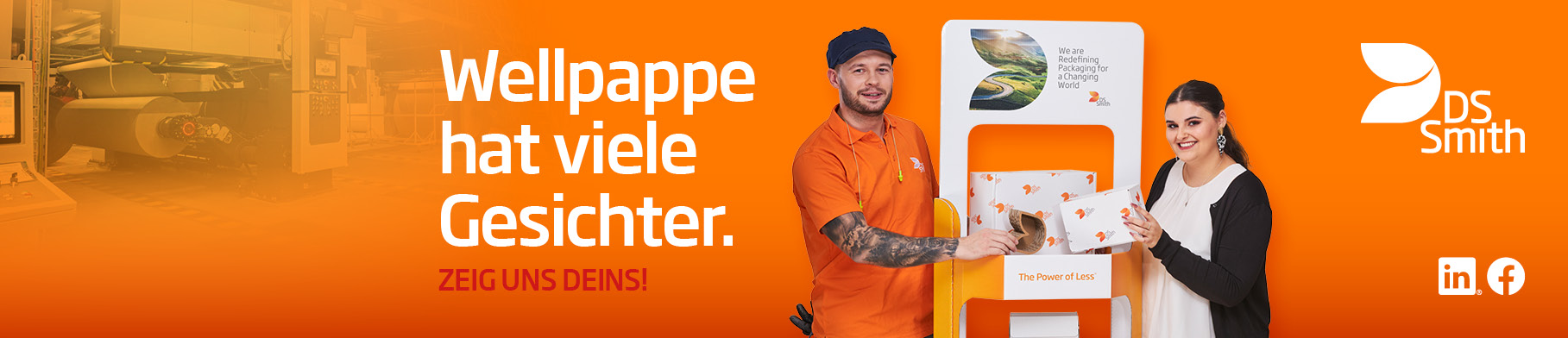 Links vom Aufsteller aus Wellpappe steht ein junger farbiger Mann mit schwarzen Haar in orangenem T-Shirt und lächelt in die Kamera. Rechts steht eine junge dunkelhaarige Frau mit weißer Bluse und dunkler Strickjacke und lächelt in die Kamera. Beide haben ein kleines Produkt aus Wellpappe in der Hand.