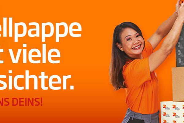 asiatische Frau mit langen Haaren steht seitlich eines Kartonstapels aus Wellpappe vor orangefarbenem Hintergrund und lächelt das Wellpappprodukt in der Hand an. Sie trägt ein orangefarbenes T-Shirt was deutlich Arbeitskleidung der Produktion ist.