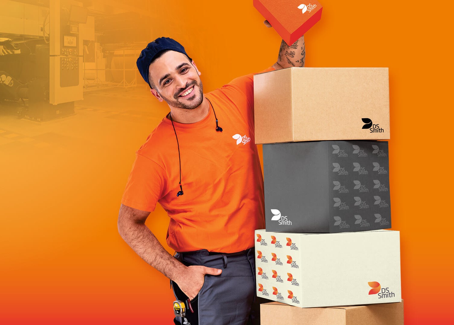 dunkelhaariger junger Mann links neben einem Kistenstapel aus Wellpappe, freundlich in die Kamera lächelnd, mit einem kleinen Wellpappkarton in der Hand. Mütze und Gehörschutz und oranges T-Shirt, Arbeitskleidung, vor orangefarbenem Hintergrund