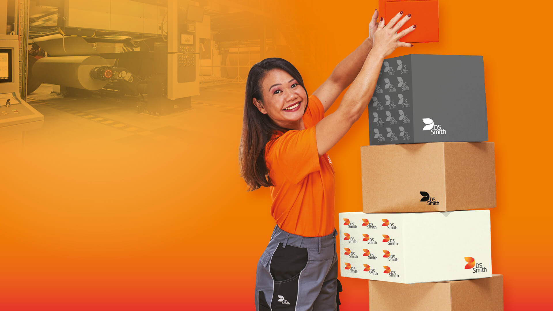asiatische Frau mit langen Haaren steht links eines Kartonstapels aus Wellpappe vor orangefarbenem Hintergrund und lächelt in die Kamera. Sie trägt ein orangefarbenes T-Shirt was deutlich Arbeitskleidung der Produktion ist. Sie hält mit beiden Händen einen kleinen Karton aus Wellpappe und stellt diesen auf den Stapel oben drauf.
