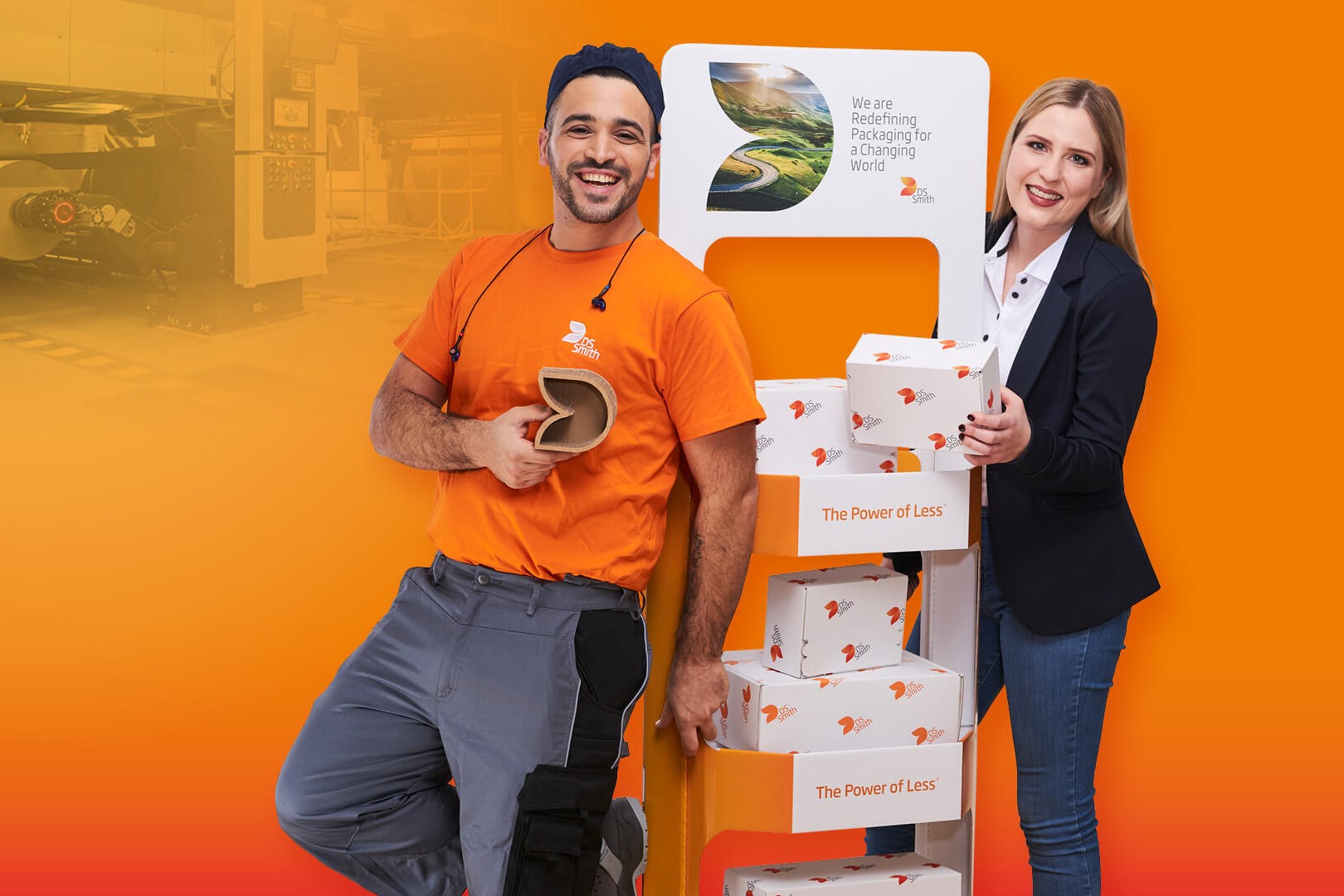 dunkelhaariger junger Mann steht links neben einem Aufsteller aus Wellpappe, freundlich in die Kamera lächelnd, mit einem kleinen Wellpappkarton in der Hand. Mütze und Gehörschutz und oranges T-Shirt, Arbeitskleidung, vor orangefarbenem Hintergrund. Rechts vom Aufsteller steht eine junge blonde Frau mit langen Haaren und dunklem Jacket und lächelt in die Kamera