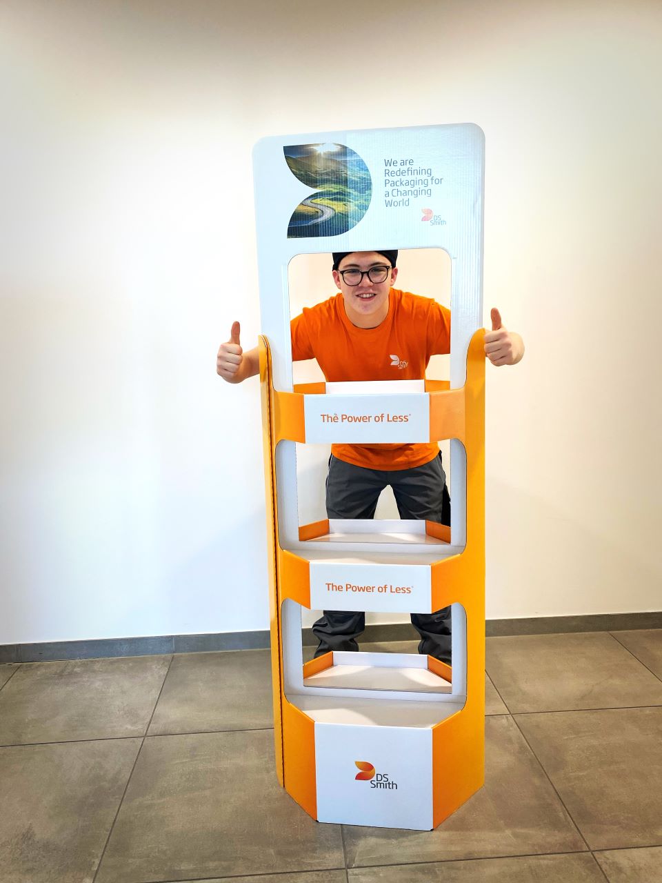 junger Mann, Mütze und dunkle Brille. Schaut durch einen Aufsteller aus Wellpappe durch, lächelt und hält beide Daumen hoch, in die Kamera.